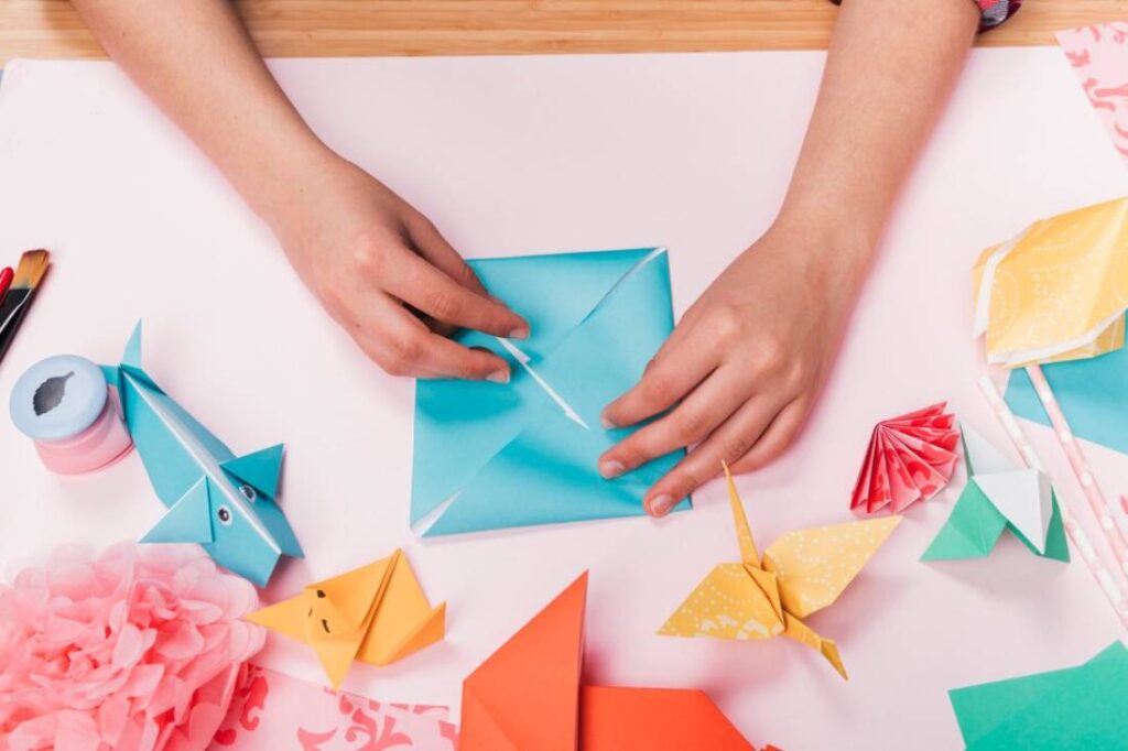 A person’s hands fold a blue origami envelope, surrounded by colorful paper creations.