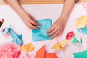 A person’s hands fold a blue origami envelope, surrounded by colorful paper creations.
