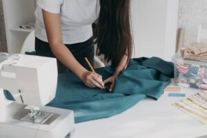 A person marking a teal fabric with a pencil at a sewing machine setup.