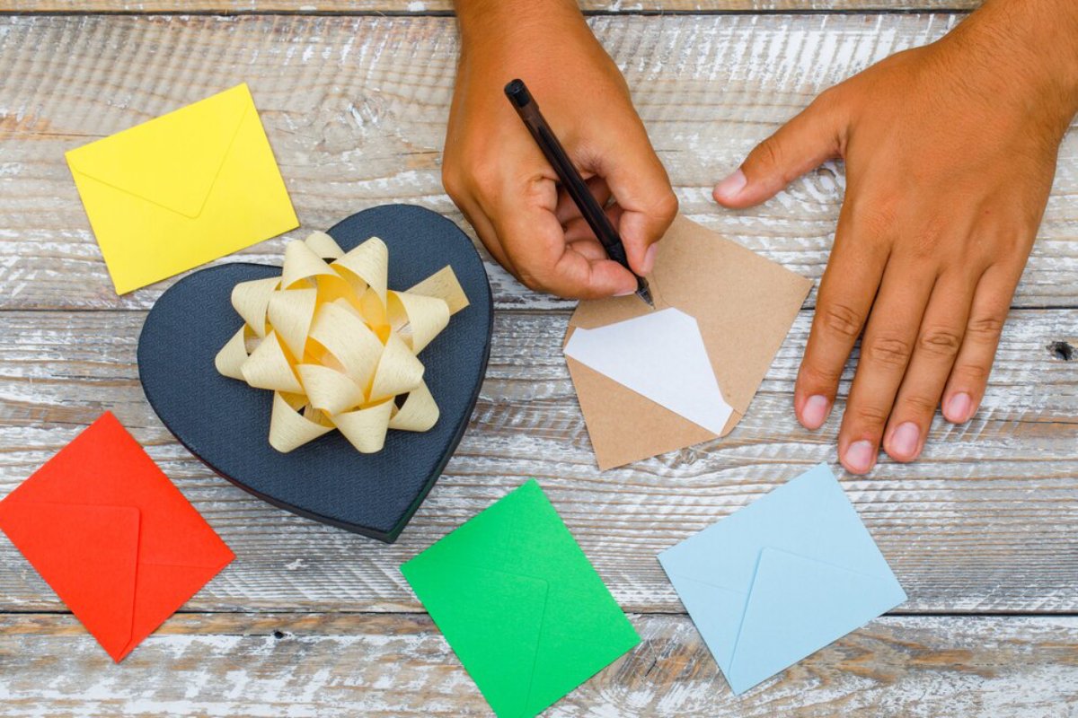 A person's hand writes on a brown envelope surrounded by colorful envelopes, a decorative ribbon, and a heart-shaped gift box.