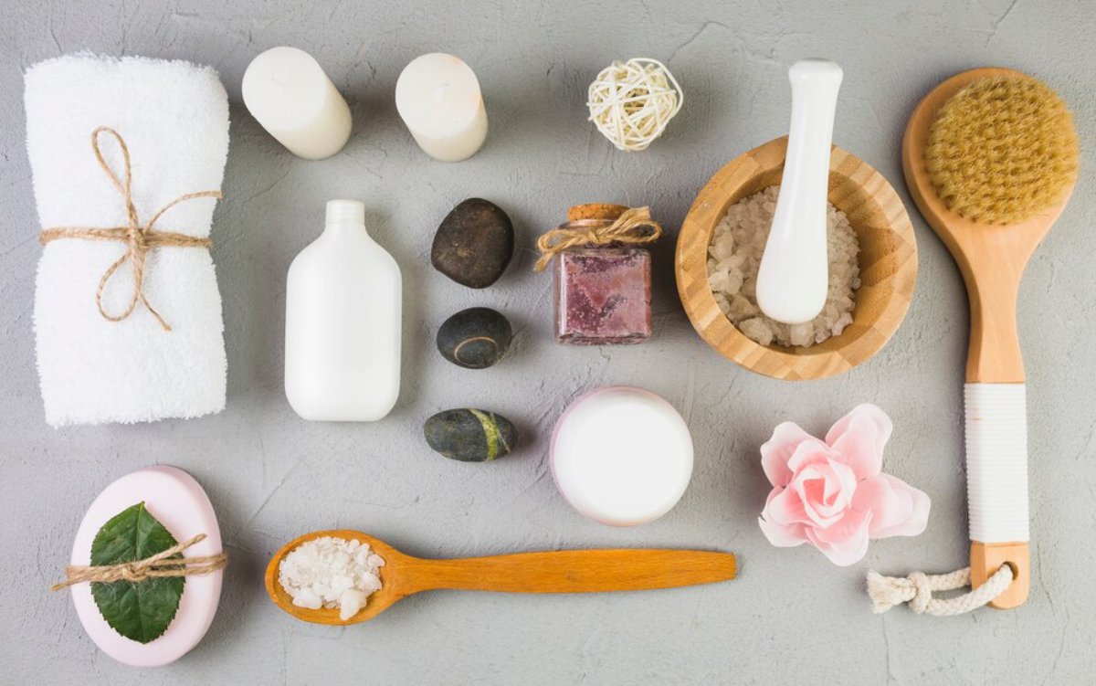An aesthetic arrangement of spa items: towels, candles, lotion bottles, stones, a brush, and decorative elements on a gray surface.