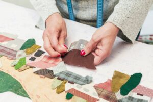 A close-up of hands sewing colorful fabric pieces resembling houses on a patterned background, demonstrating textile craft skills.