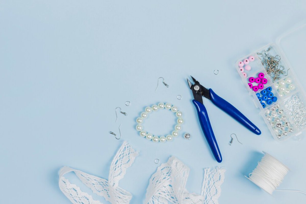 Crafting tools and materials arranged on a light blue background, including pearls, lace, colorful beads, and jewelry pliers.