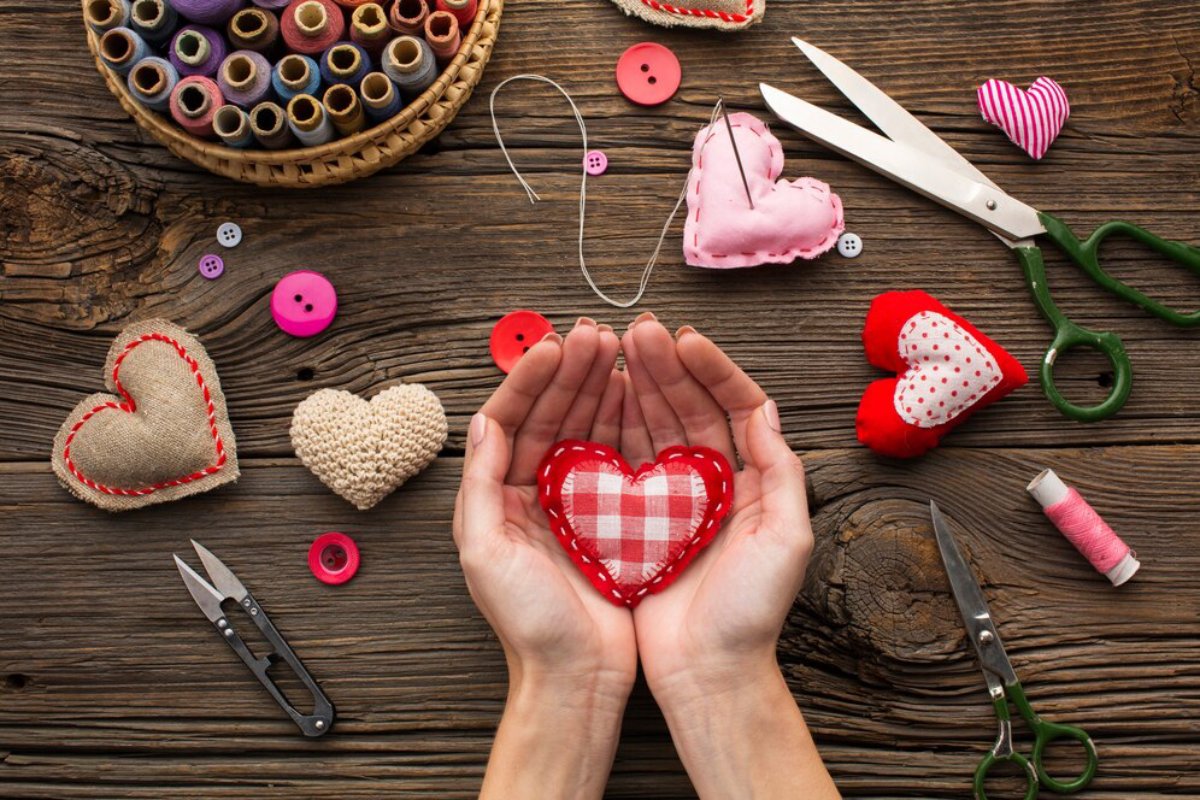 A pair of hands cradles a stitched red heart, surrounded by buttons, thread spools, and various heart-shaped fabric crafts on wood.