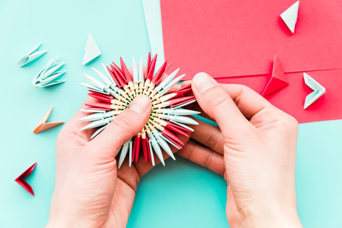 A person holding a colorful origami piece surrounded by various folded paper shapes on a light blue background.