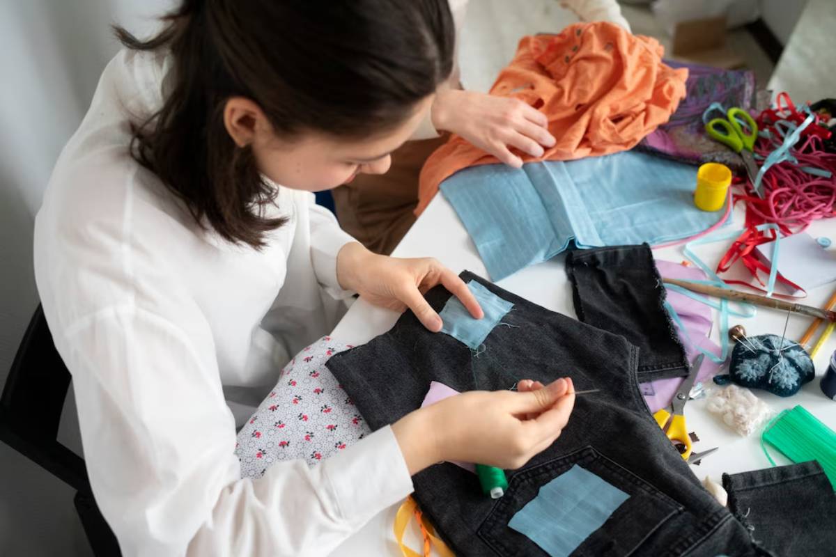 A person is hand-sewing colorful patches onto dark fabric, surrounded by various sewing supplies and a vibrant array of materials.