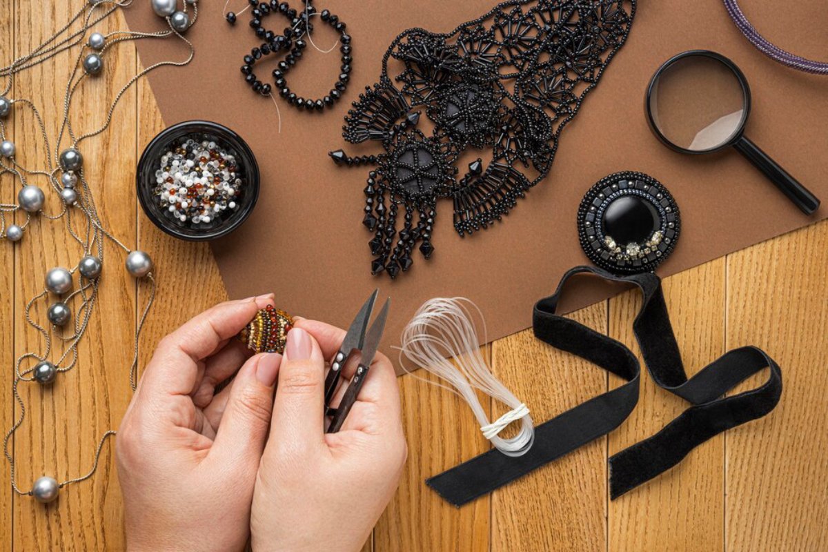 A pair of hands holds colorful beads and scissors, surrounded by various crafting materials including a magnifying glass and black lace.