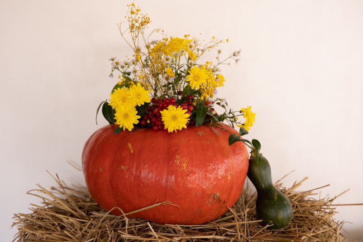 A large orange pumpkin serves as a vase, adorned with yellow flowers and greenery, placed atop a straw base.