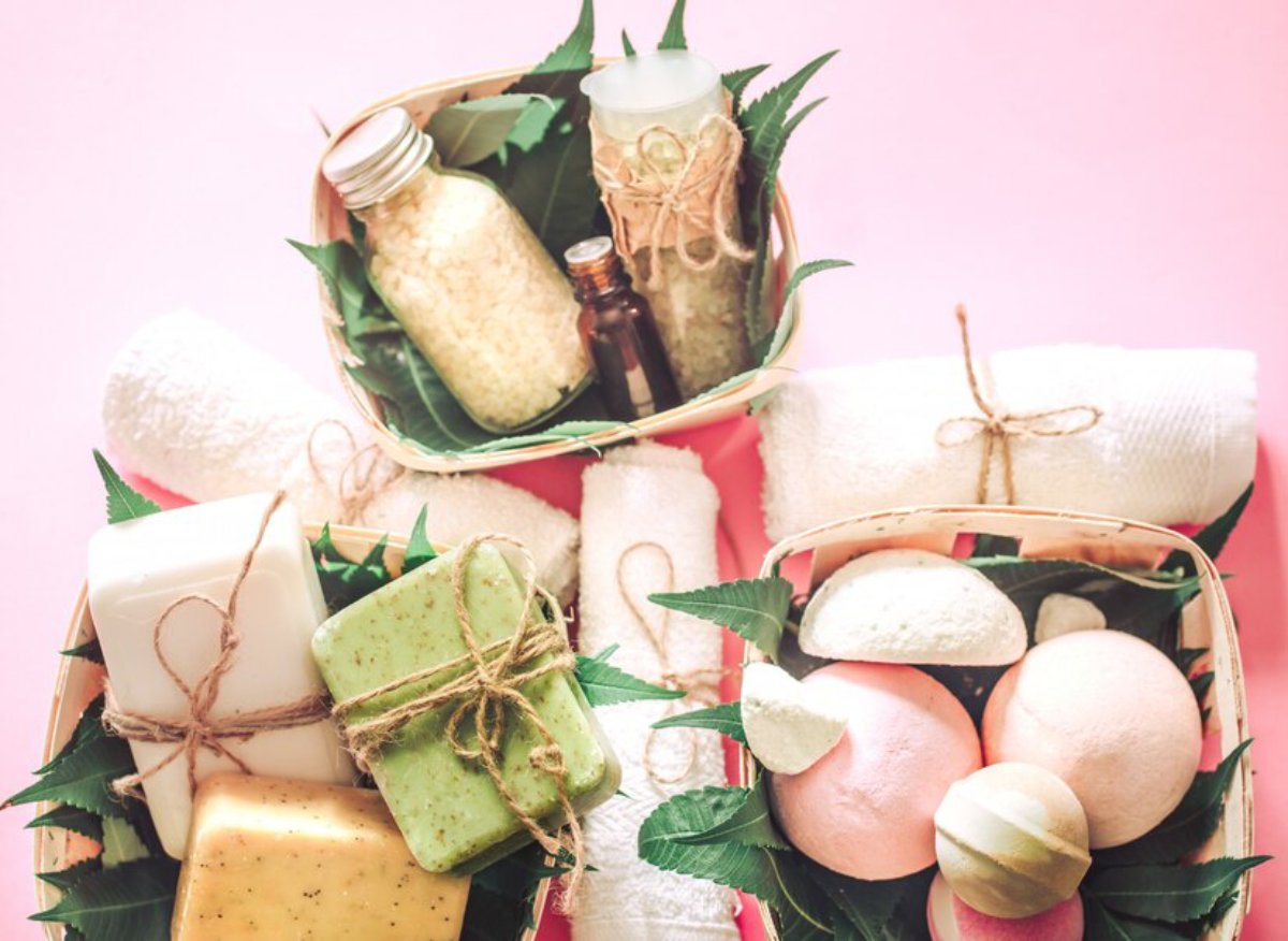 Two woven baskets filled with colorful soaps, bath bombs, and natural products surrounded by leafy greenery on a pink background.