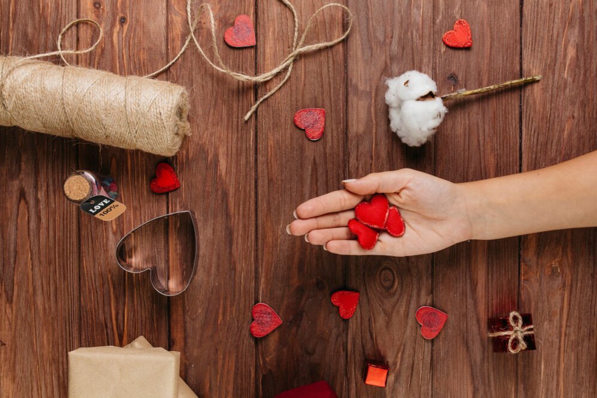 A hand holds three small red hearts, surrounded by twine, cotton, gift wrap, and more heart-shaped decorations on a wooden surface.