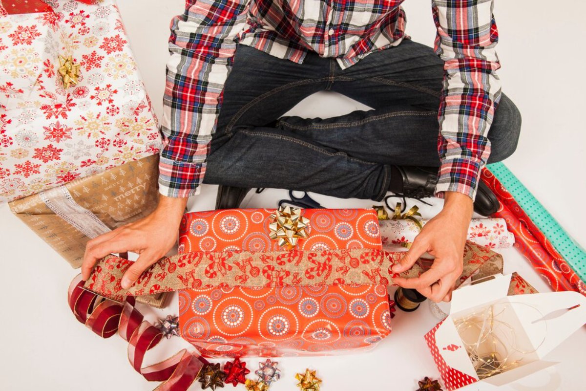 A person in a plaid shirt wraps a festive orange gift, surrounded by colorful wrapping paper, ribbons, and decorative bows.