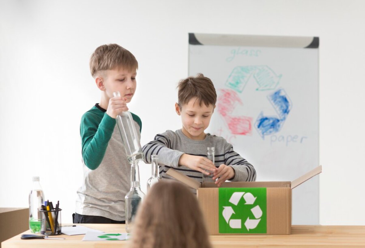 Two children are engaged in a recycling activity, sorting bottles into a cardboard box marked with a recycling symbol.