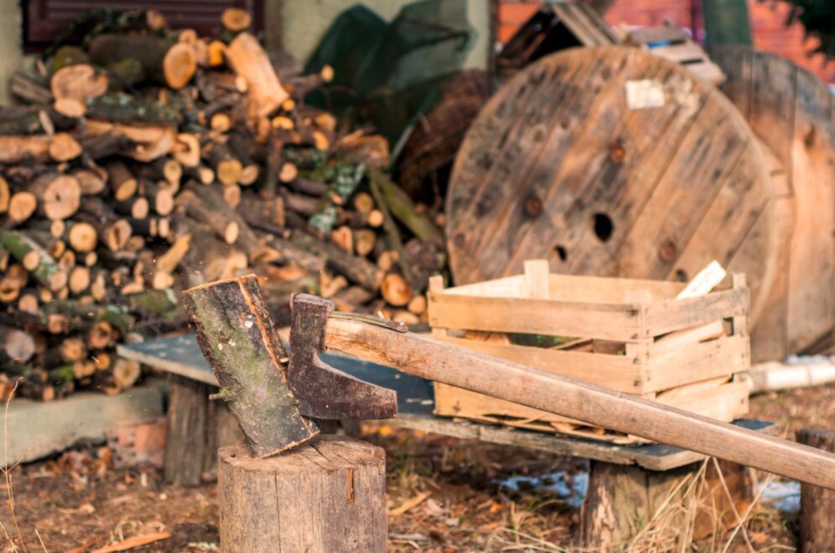 An axe embedded in a wooden stump, with a backdrop of stacked firewood and a wooden crate in a rustic outdoor setting.