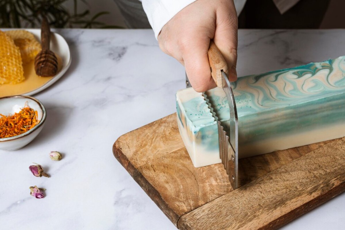A hand holds a wooden knife slicing a marbled soap bar on a cutting board, with honeycomb and dried flowers nearby.