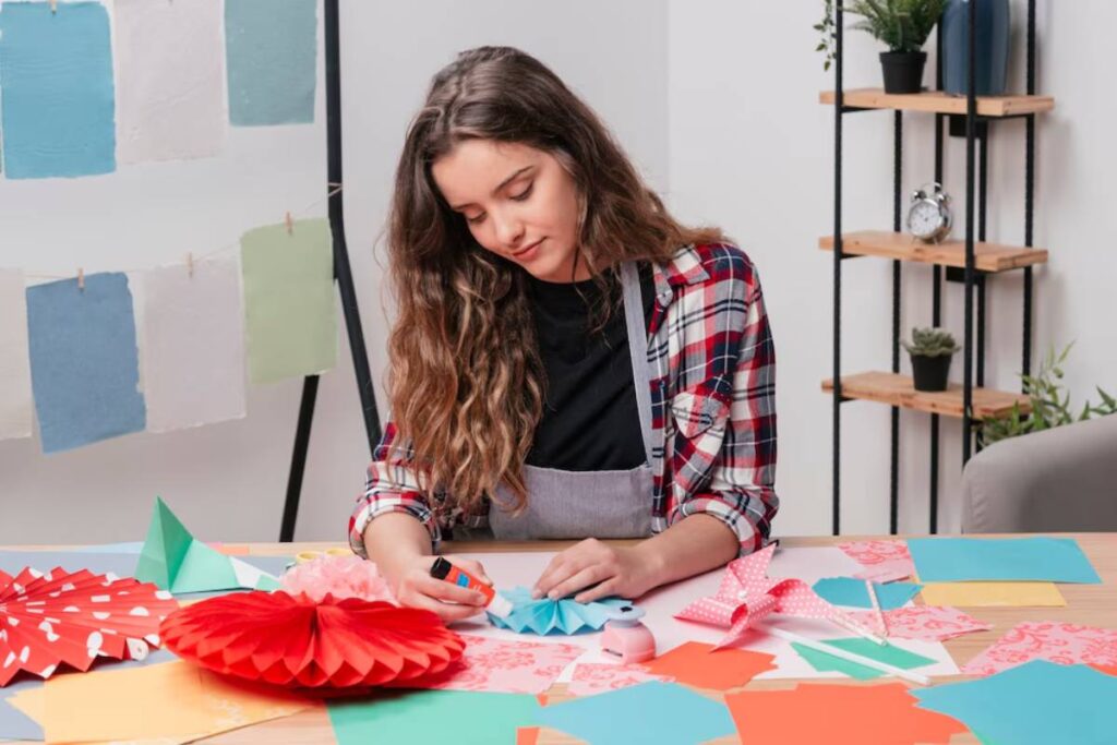 A person is crafting with colorful paper and glue, surrounded by various shapes and decorations in a bright workspace.