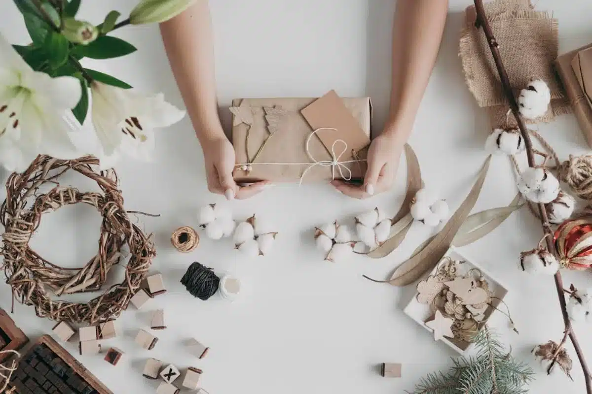 A person holds a wrapped gift surrounded by crafting materials, cotton, and decorative elements on a white table.