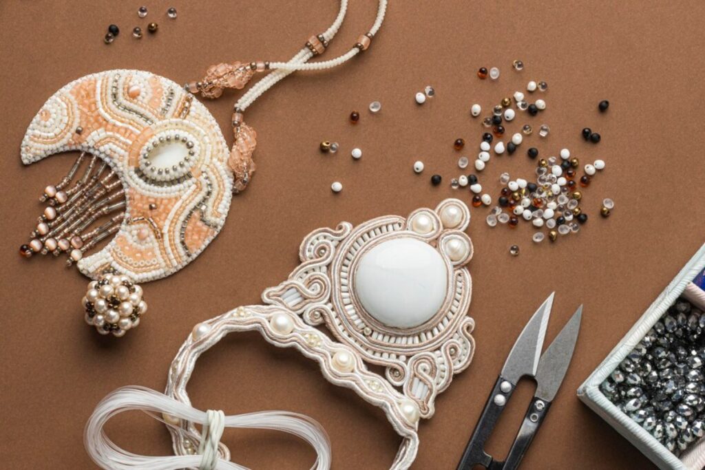 Beaded jewelry pieces and beads scattered on a brown surface, with scissors and a box of rhinestones nearby.