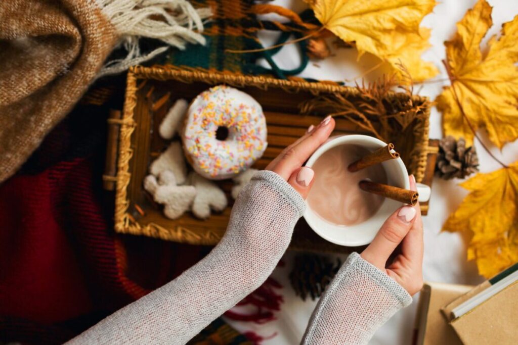 A cozy scene featuring a hand holding a mug of hot chocolate with cinnamon sticks, a sprinkled donut, and autumn leaves in the background.