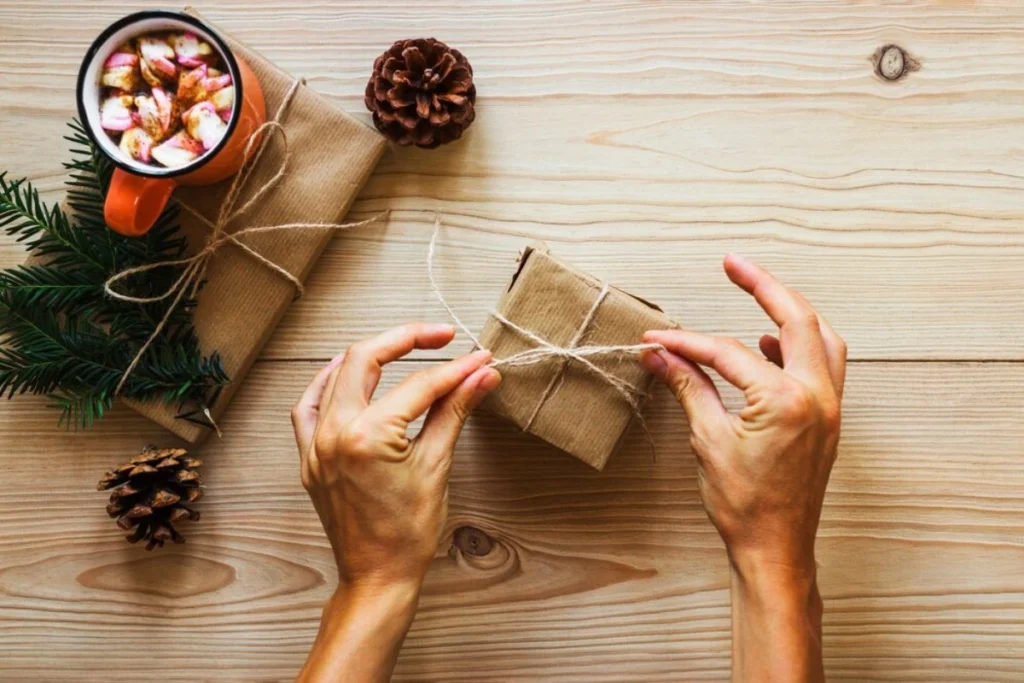 A person wraps a small gift box in brown paper and twine, surrounded by holiday decor and a mug of hot chocolate with marshmallows.
