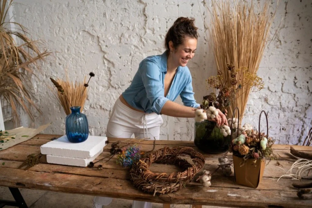 A woman in a blue shirt arranges flowers and dried plants on a wooden table, surrounded by vases and creative decor items.